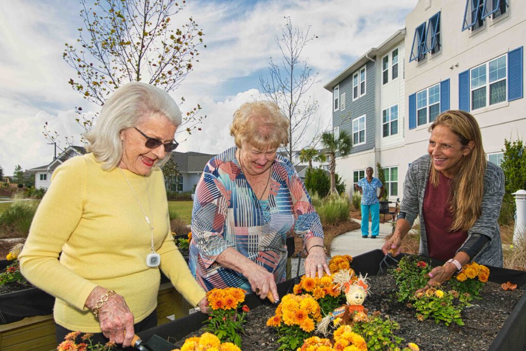 Three older white women gardening outdoors and smiling as they're working on orange flowers at Portside at Grande Dunes, a senior living community near Conway, SC.