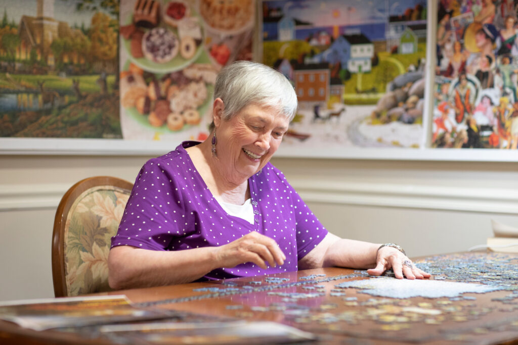 Grace_Management_Memphis_2018-11 - Portside at Grande Dunes An older white woman wearing a purple dress with small white polka dots is smiling as she's sitting at the table and putting together a puzzle at Portside at Grande Dunes, a senior living community near Murrells Inlet, SC.