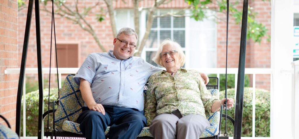 Grace_Management_Memphis_2018-5362 - Portside at Grande Dunes A middle-aged white man is sitting on the swing next to an older white woman with his hand on her shoulder at Portside at Grande Dunes, a senior living community near Conway, SC.