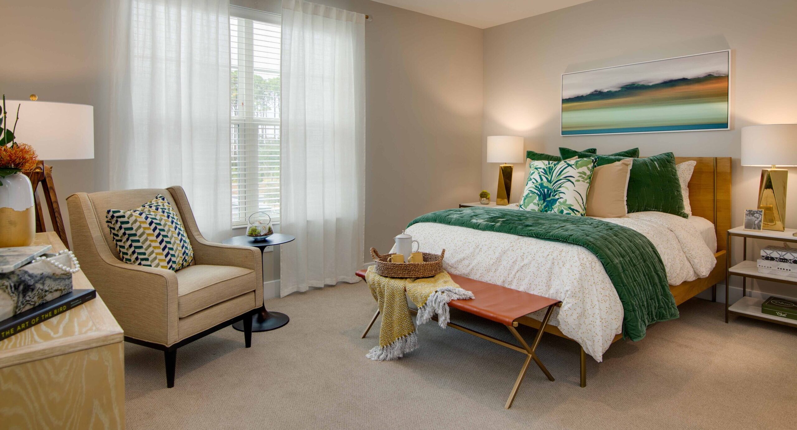 IL Model Unit 4 - Portside at Grande Dunes A bedroom with a made-up bed with white sheets, green throw blanket, green throw pillows, a landscape painting above the bed, two bedside tables with lamps on each side, a sitting sofa chair across the room, and an orange footstool with a yellow throw blanket on it. Portside at Grande Dunes, a senior living community near Murrells Inlet, SC.