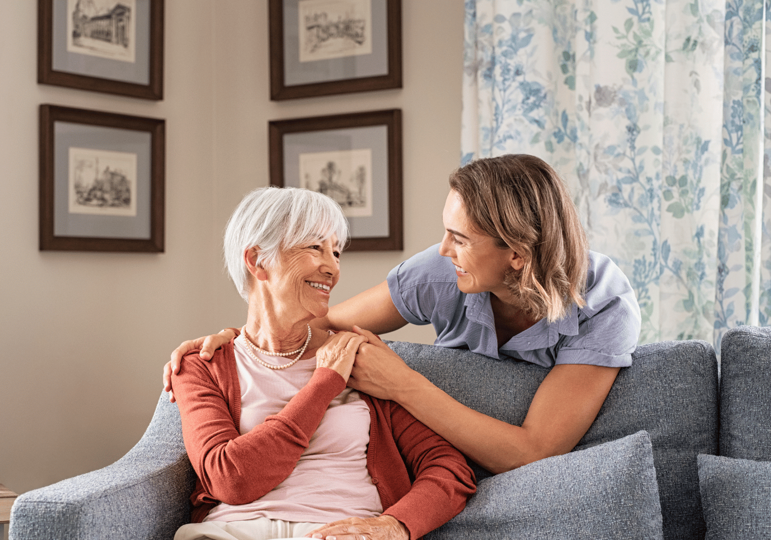 Smiling elderly woman with white hair on a grey sofa, wearing a pink top and red cardigan, being comforted by a younger caregiver.