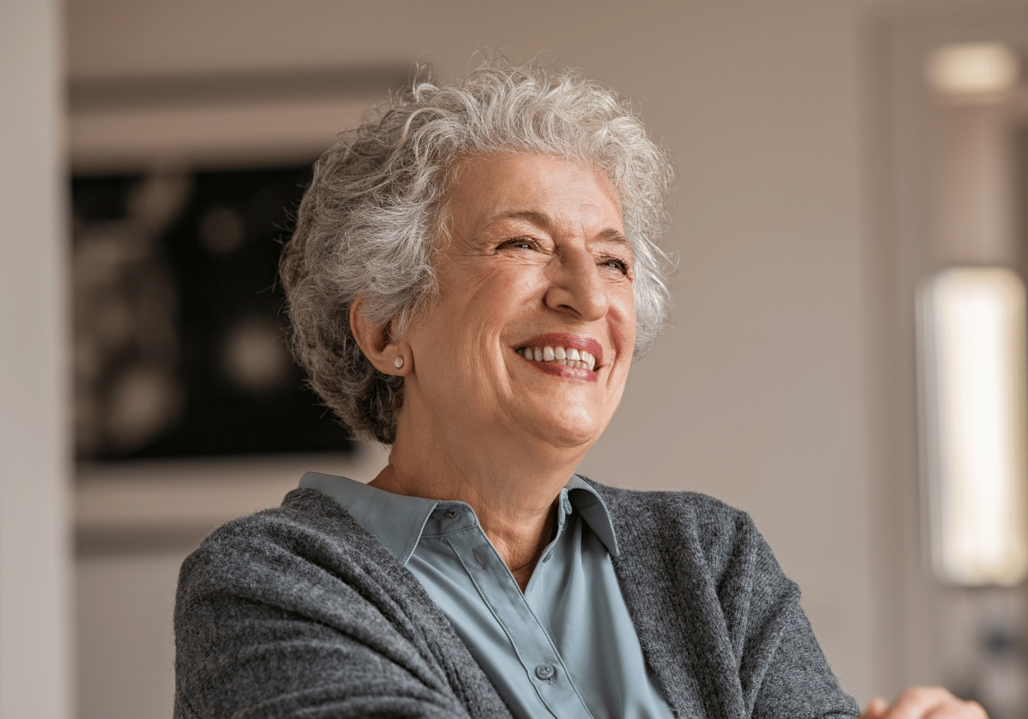 Smiling elderly woman with gray hair wearing a blue blouse and gray cardigan, indoors.